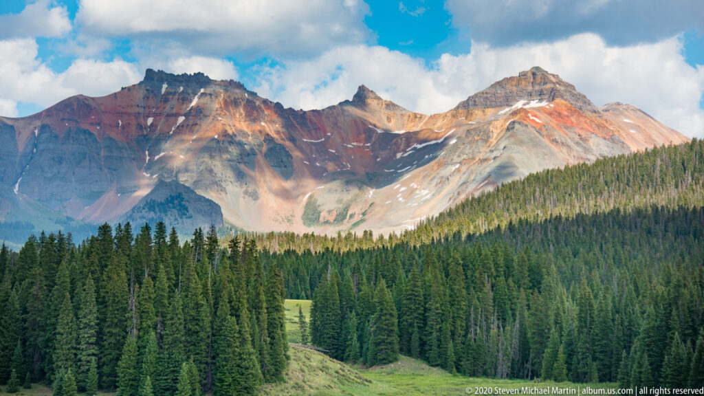 A lake fills a forested valley beneath distant alpine peaks, with meadows and small buildings along shore.