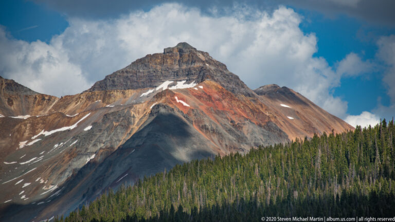 A red-brown alpine summit with small snow patches rises above a dark conifer forest.