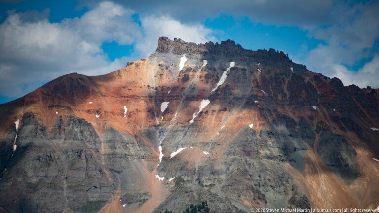 Red, orange, and gray rock bands descend from a dark crest, with snow in several gullies.