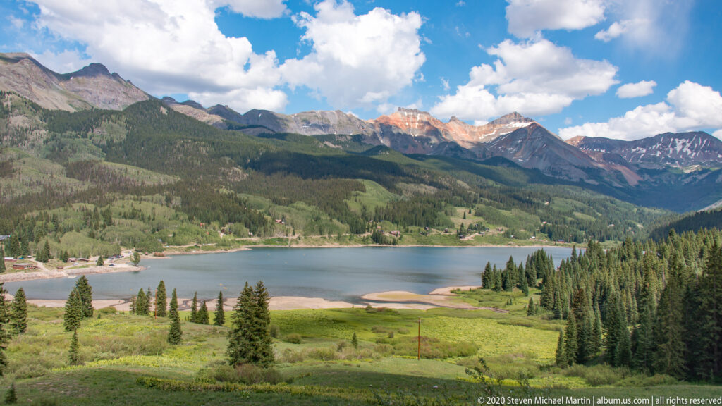 A lake sits below forested ridges and snow-dotted peaks, with green meadows descending to shore.
