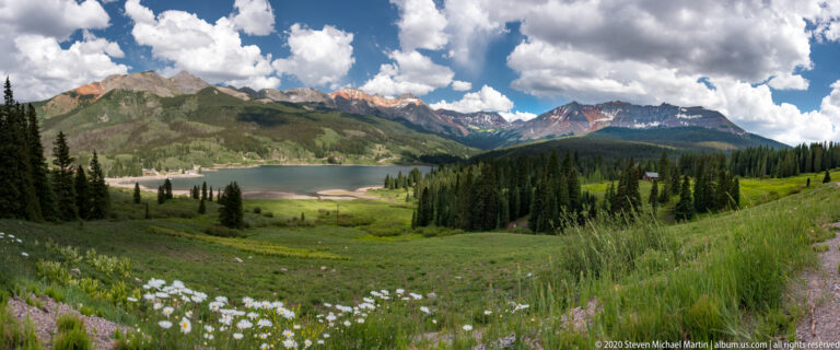 Flowers and meadow plants overlook a lake bordered by conifers and colorful alpine peaks.