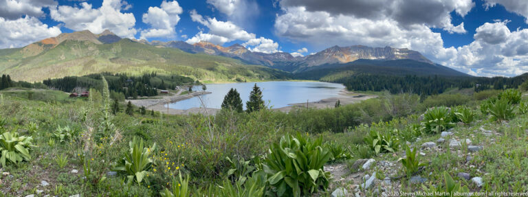 A mountain lake lies below forested slopes and high peaks, with small buildings along the shore.