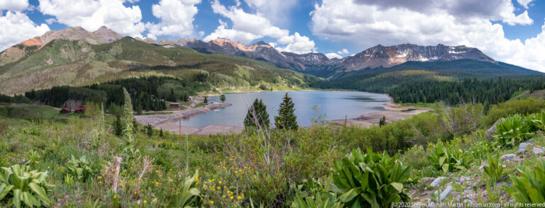 A flowered hillside descends to a lake bordered by forest, with high peaks beyond.