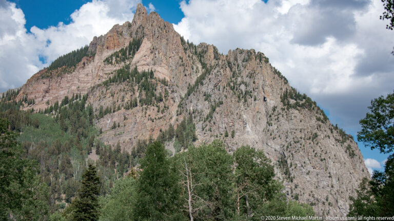 A jagged rocky wall rises above conifers and lighter trees beneath broken clouds.
