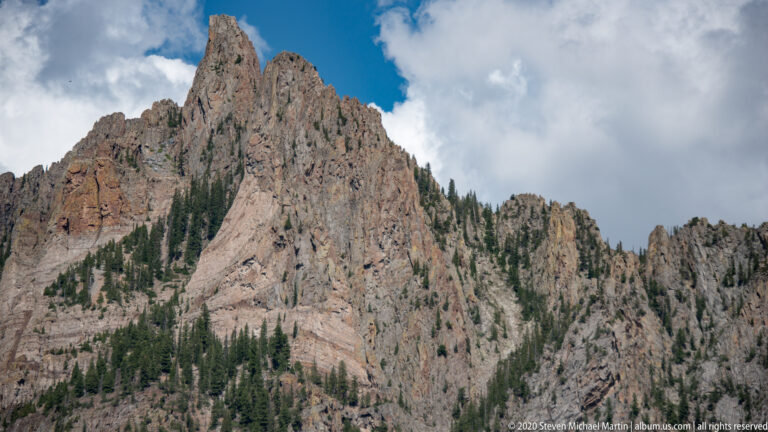 A jagged gray-brown summit rises above forested lower slopes beneath gathering clouds.
