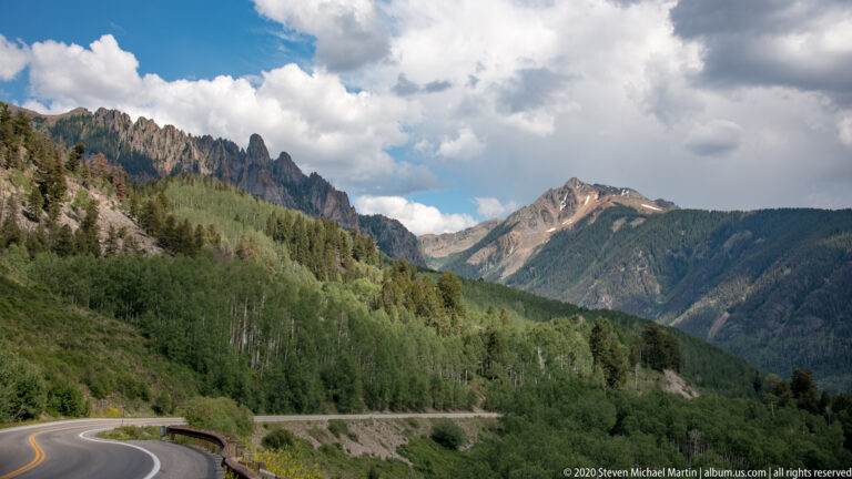 A paved road curves below forested slopes and jagged, snow-patched mountain peaks.