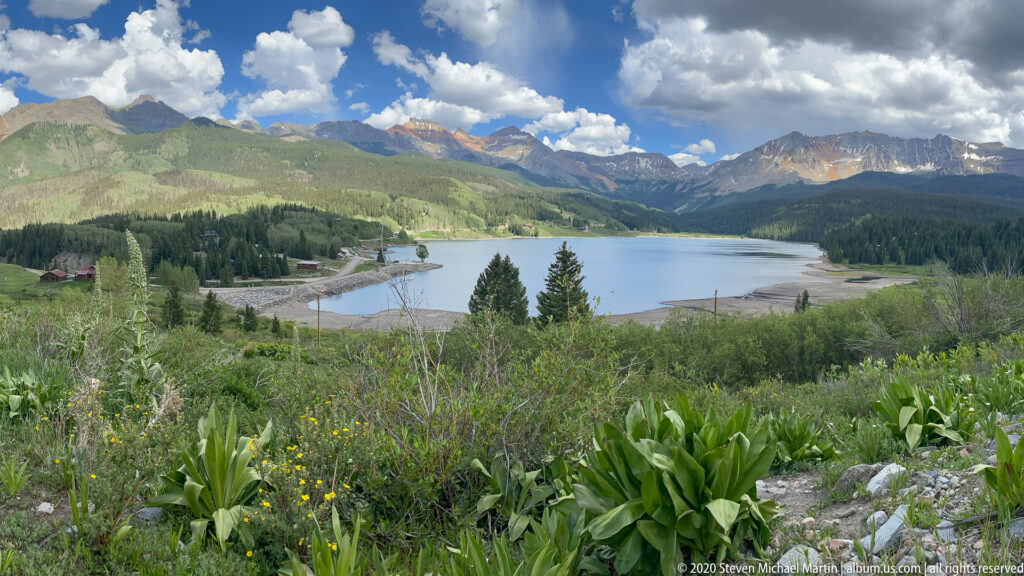 Meadow plants frame a mountain lake and small shore buildings beneath a rugged peak skyline.