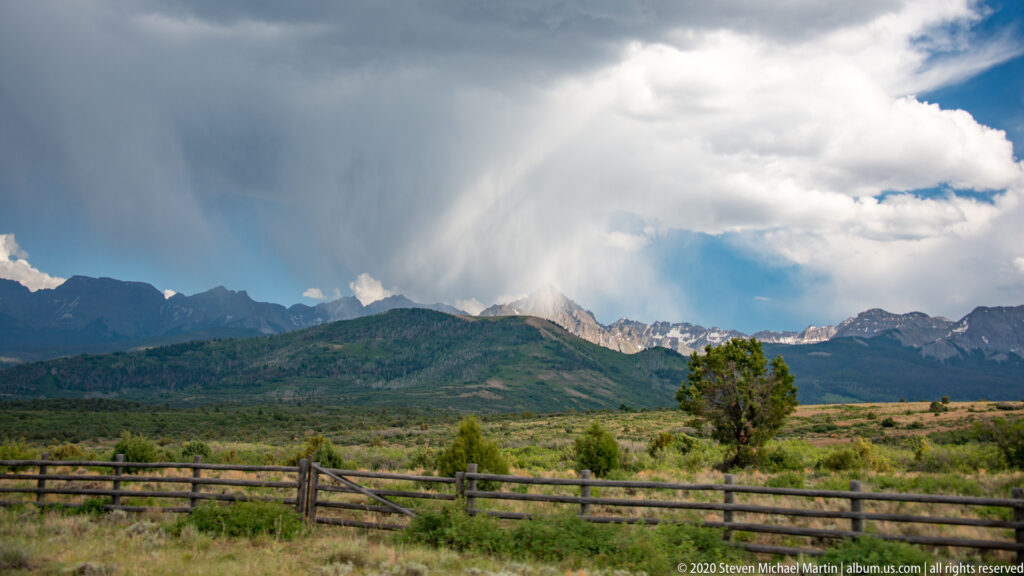 A towering storm cloud and rain shaft loom over jagged mountains beyond a rustic fence.