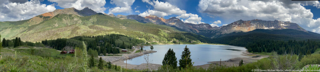 Tall meadow plants overlook a lake, cabins, forested slopes, and jagged alpine peaks.