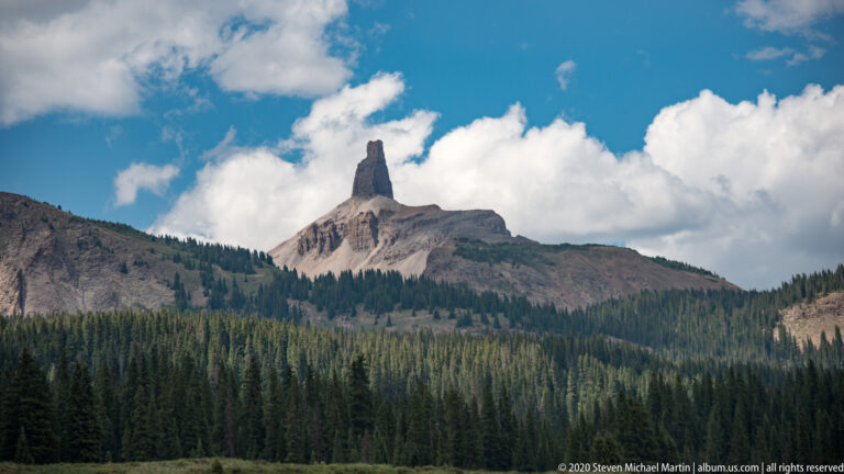 A narrow rock spire projects from a pale ridge above a dense evergreen forest.
