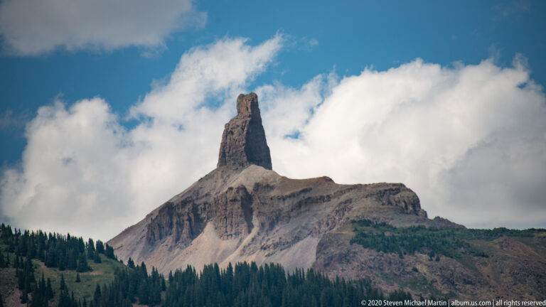 A dark rock pinnacle rises from a pale summit shoulder above a band of conifer forest.