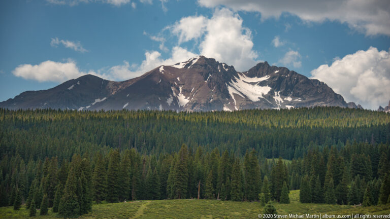 Snow patches mark dark mountain slopes above a dense evergreen forest and open meadow.