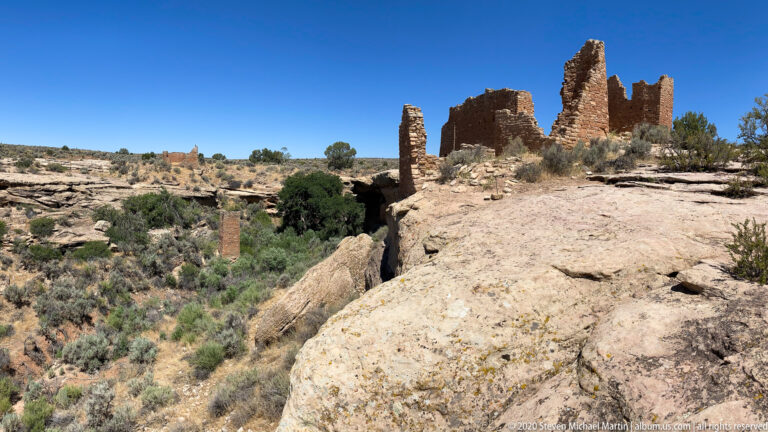 Stone ruins stand on opposite sides of a narrow canyon beyond a broad sandstone ledge.