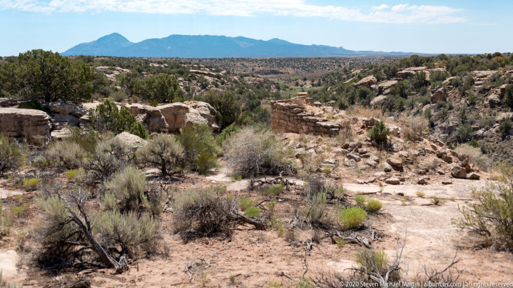 Scattered masonry ruins sit among shrubs and junipers across a broad rocky plateau.