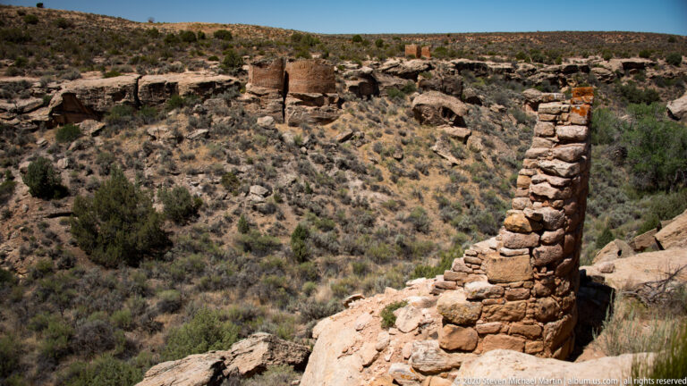 A narrow stone tower rises at a canyon edge above scattered ruins and desert shrubs.
