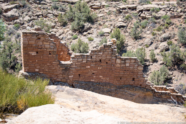 A long stepped masonry wall with a dark opening stands against pale rock and desert shrubs.