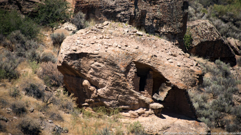 A large sandstone boulder overhangs a masonry house with a dark recessed doorway.