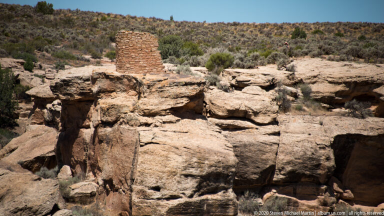 A small stone tower stands above a dark recess beneath a broad sandstone shelf.