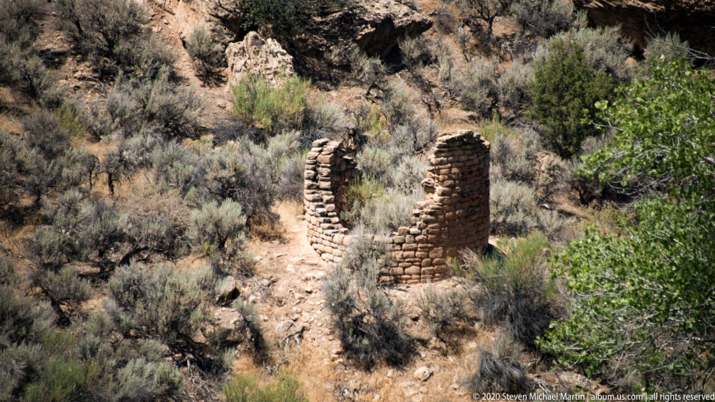 Two rounded masonry walls stand apart among sagebrush and juniper on a rocky slope.