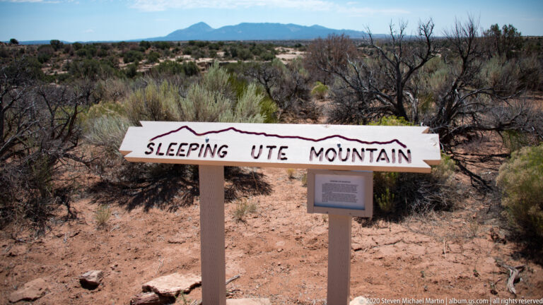 A white sign reading “Sleeping Ute Mountain” stands above a smaller placard in dry scrub.