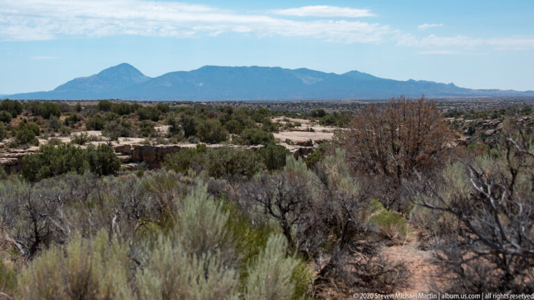A blue mountain range spans the horizon beyond sagebrush, junipers, and exposed rock.