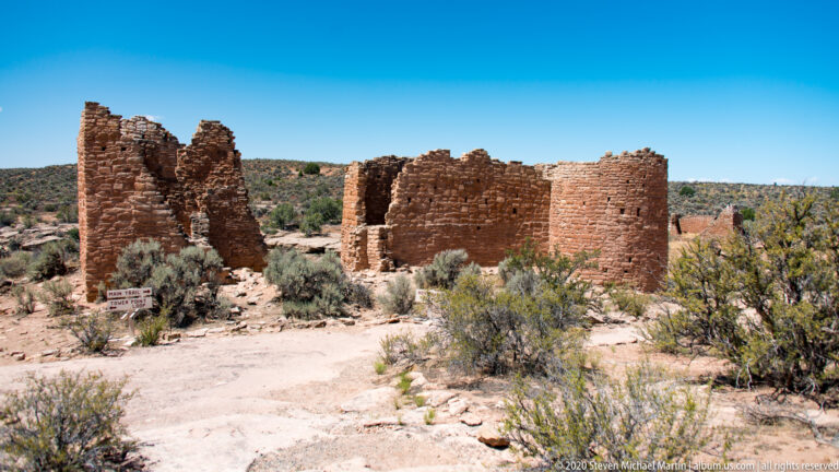 Hovenweep Castle stands among shrubs above a sandy path, with mesas visible beyond.