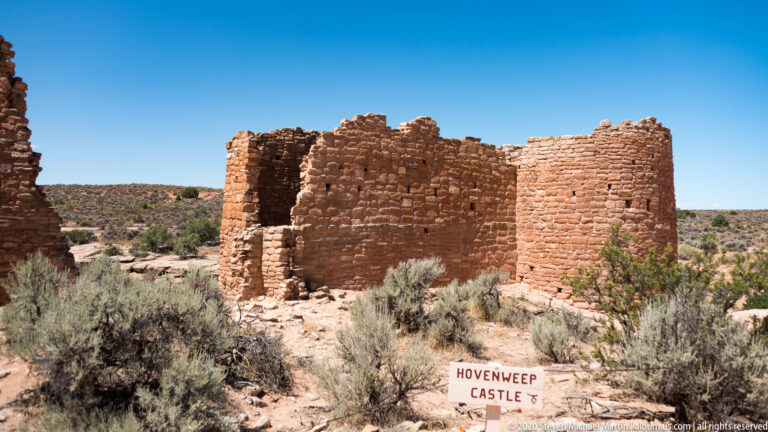 A stone ruin with a rounded tower and dark opening stands behind a sign reading “Hovenweep Castle.”
