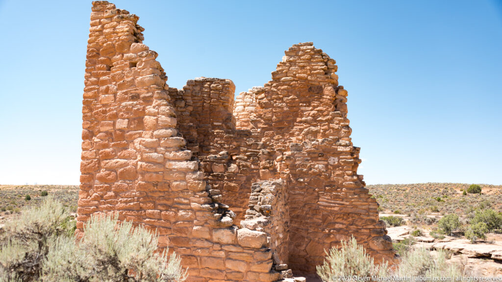 Two tall sandstone wall remnants rise above a lower section against a clear blue sky.