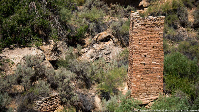 A rectangular stone tower stands on a rocky ledge above a shrub-filled ravine with lower wall fragments nearby.