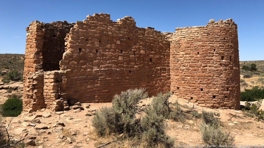A broad sandstone masonry ruin with a dark opening stands above scrub and scattered stones.