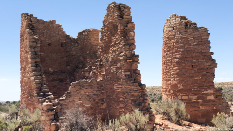Two tall stone ruin walls with dark openings rise above low masonry and desert brush.