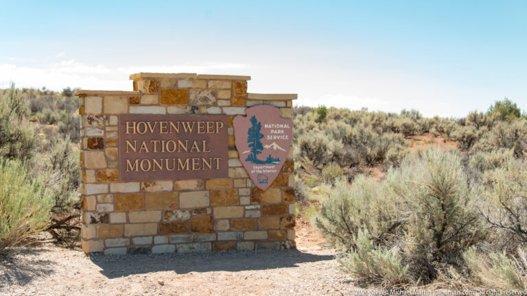 A stone sign reads “Hovenweep National Monument” beside an NPS arrowhead in desert scrub.