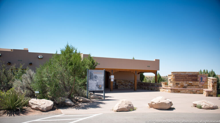 A low visitor center and stone sign stand beyond a paved approach bordered by boulders and shrubs.