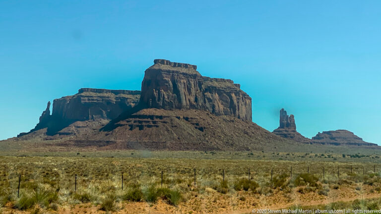 A tall flat-topped butte and slender spire rise above a fenced desert plain.