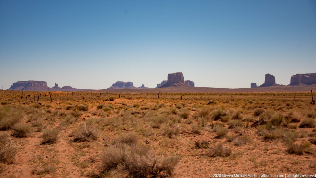 A broad desert plain and faint track lead toward a distant line of buttes and spires.