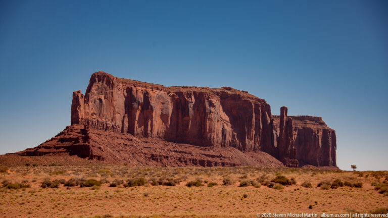 A long red cliff with a detached slender spire rises above a scrub plain.