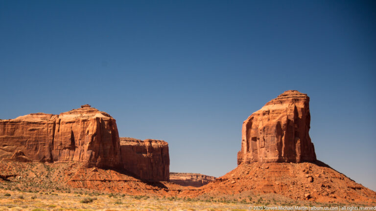 Two red sandstone formations stand apart across a scrub plain, with distant mesas in the gap.