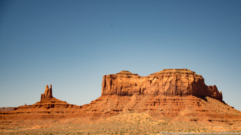 Narrow pinnacles and a broad mesa rise beyond a flat, shrub-covered plain.