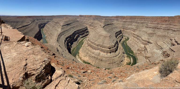 The green San Juan River curves around a striped rock promontory deep below the canyon rim.