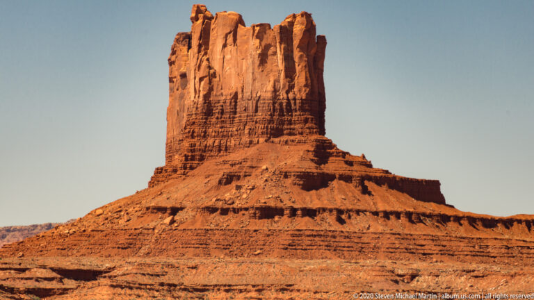 A close red butte has broken summit pinnacles above a broad stepped base.