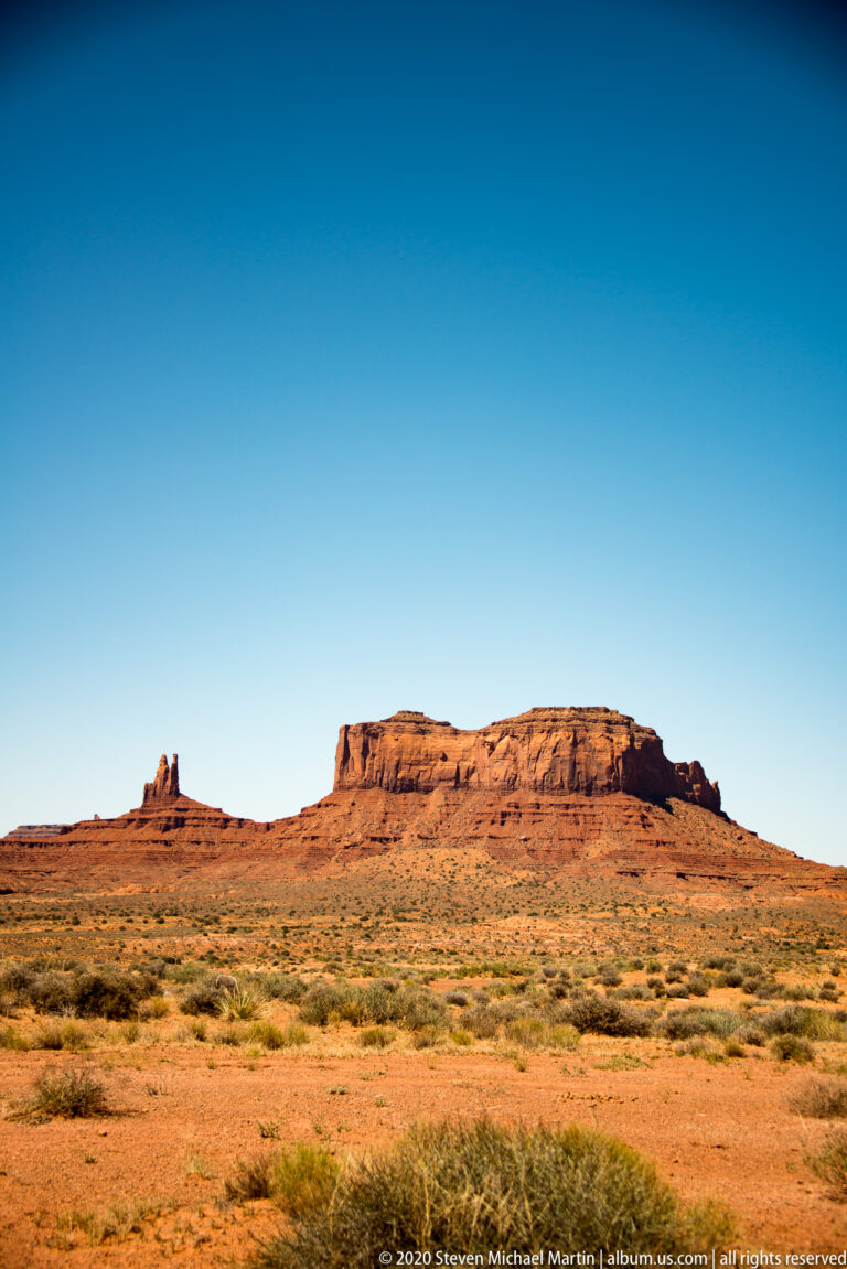 A low mesa and two narrow spires rise above a grassy desert plain.