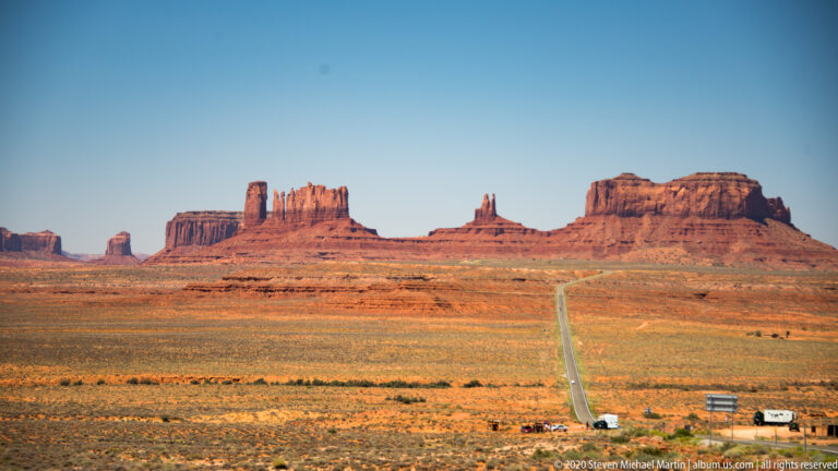 A narrow road and small vehicles lead across a plain toward a wide skyline of red buttes.