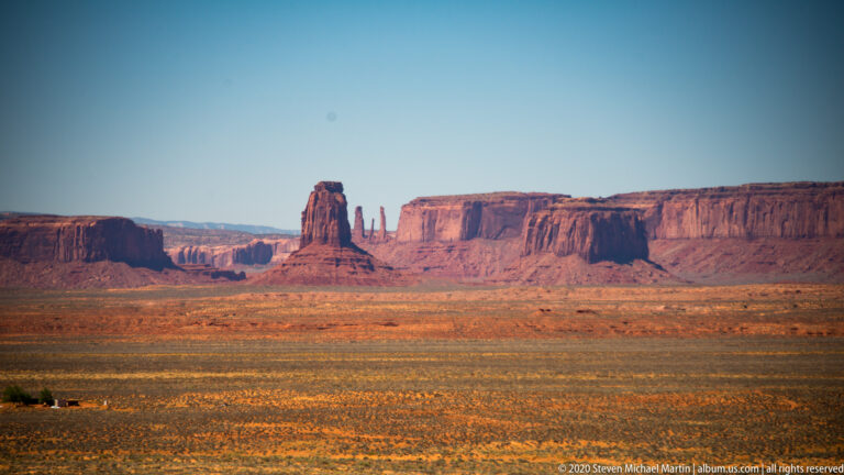 A solitary tower-shaped butte stands between distant cliffs beyond a golden scrub plain.