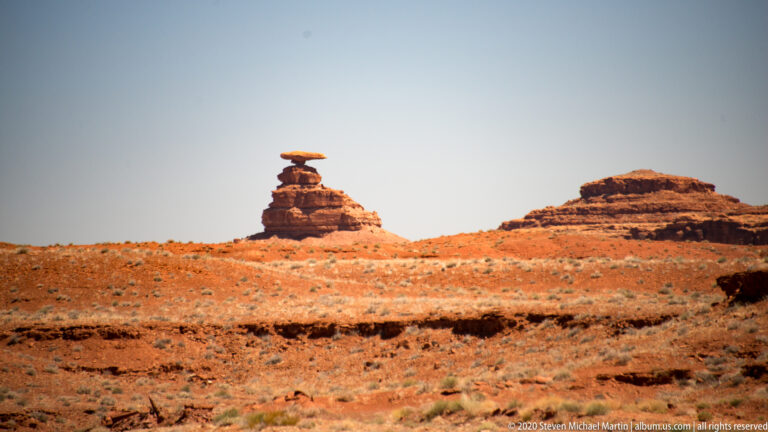 A flat dark capstone balances on a narrow red rock column above a low ridge.