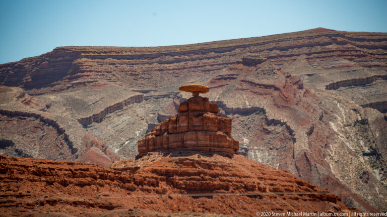 A capstone-topped rock pedestal stands before a broad layered canyon wall behind it.