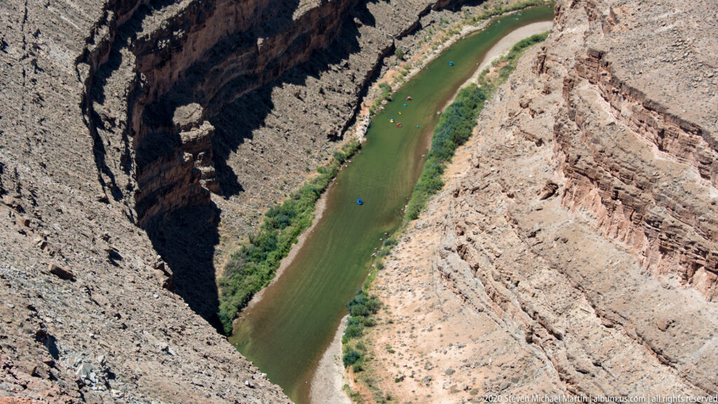 A green river runs between steep canyon walls, with tiny colorful craft on the water.