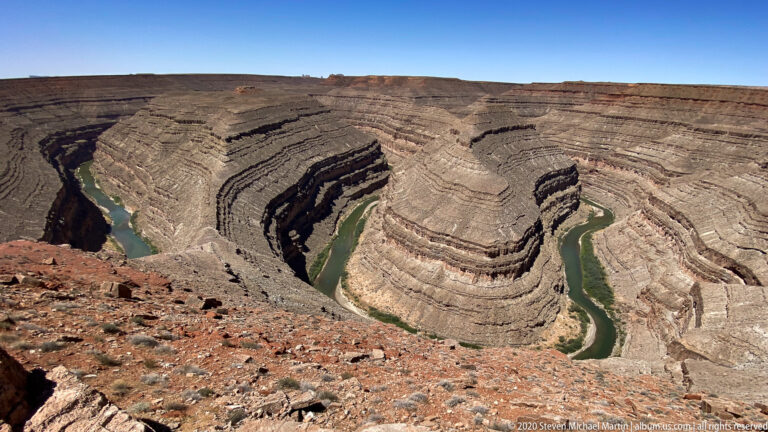 Layered canyon walls surround a green river looping tightly around a large rock spur.