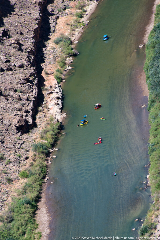 Several colorful kayaks move along a broad green river bordered by pale rock and scrub.