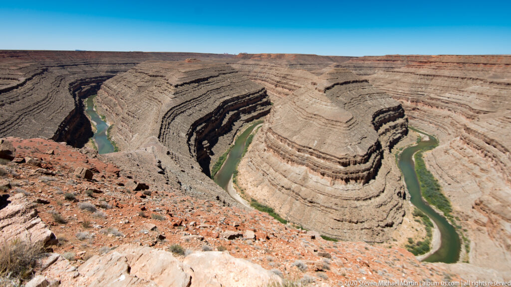 A green river winds around a broad rock mass far below a rocky canyon rim.