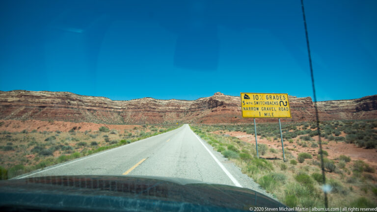 A yellow sign warns drivers of a 10 percent grade, switchbacks, and a narrow gravel road ahead.